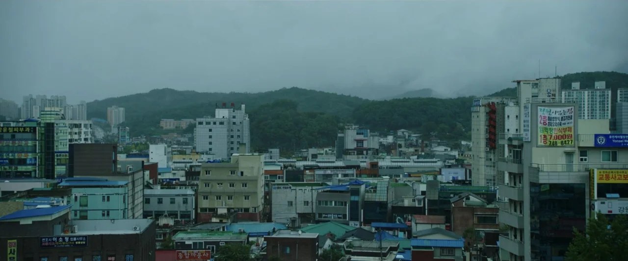 The Gangster, the Cop, the Devil (2019) movie still: establishing — A dense urban cityscape with multi-story concrete buildings beneath a thick,…