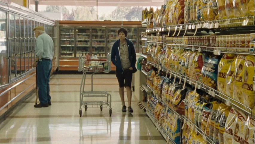 Wendy and Lucy (2008) movie still: fullbody — A quiet grocery store aisle features long shelving units filled with snack bags. An elderly man looks…