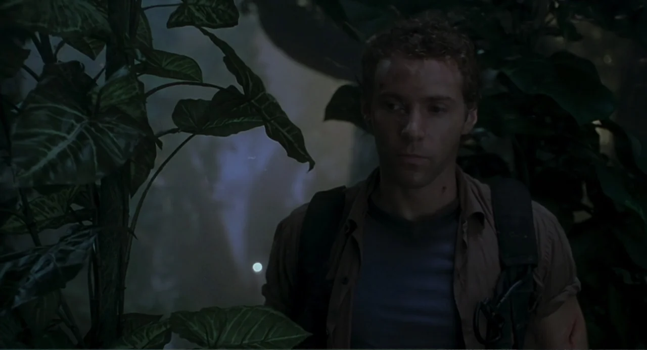 Jurassic Park III (2001) movie still: medium — A dense, overgrown tropical environment with large leaves obscuring part of the view. A young man in…