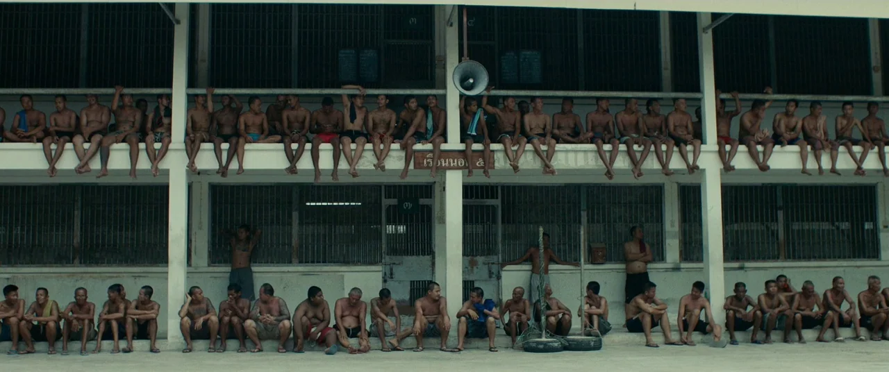 A Prayer Before Dawn (2017) movie still: wide — A long, multi-level concrete building has iron bars covering large open windows. Dozens of shirtless…