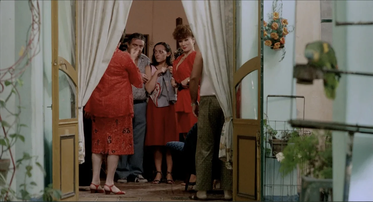 Waiting for the Hearse (1985) movie still: fullbody — A group of five people stand within a domestic doorway leading into a room. One woman in red…