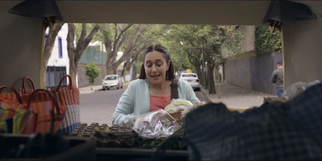 El sabor de la Navidad (2023) movie still: medium — A woman stands behind an open car trunk on a quiet tree-lined street. She sorts through grocery…