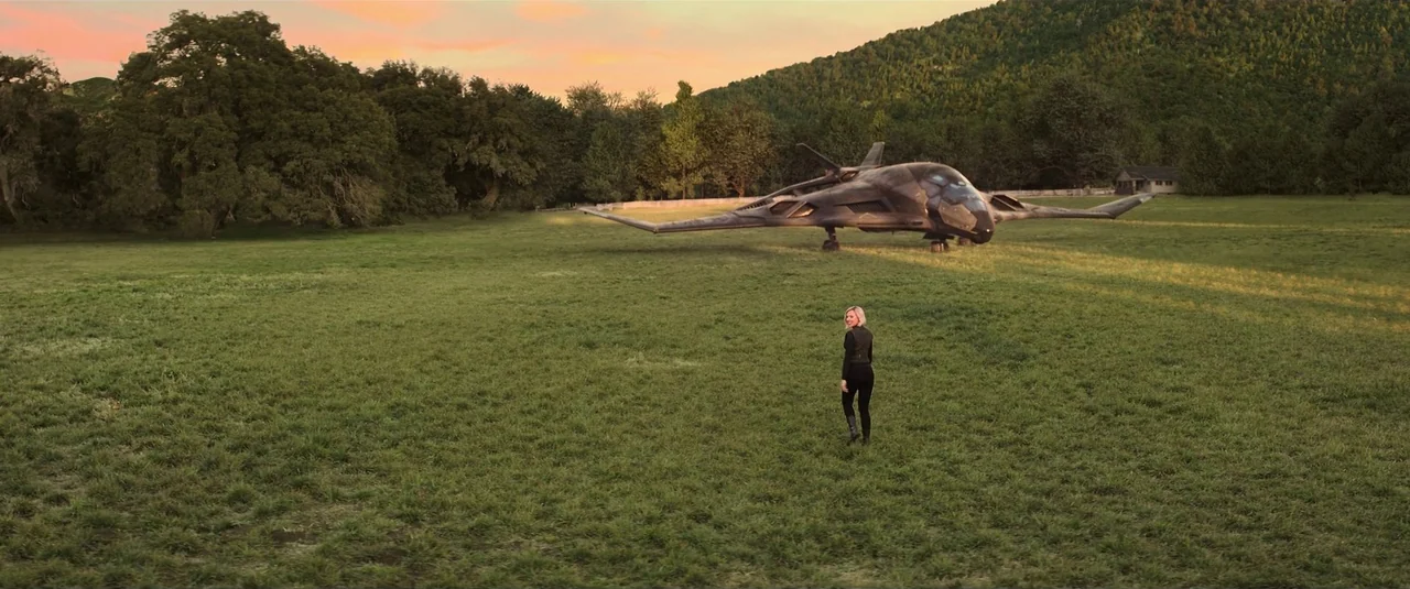 Black Widow (2021) movie still: wide — A vast grassy meadow bordered by a dense forest under a warm evening sky. A sleek, dark futuristic aircraft…