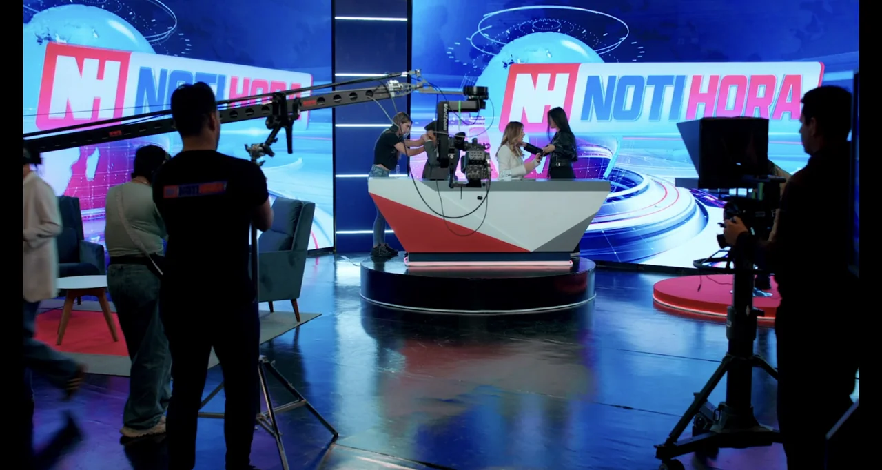Paired Up (2019) movie still: wide — A television news studio features large glowing blue screens with a central white desk. Camera operators and…