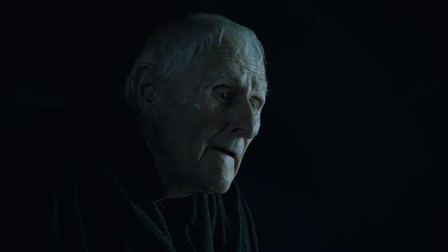 Game of Thrones (2015) movie still: closeup — A dark, nondescript interior space surrounds the elderly man in the foreground. He stands in shadows…