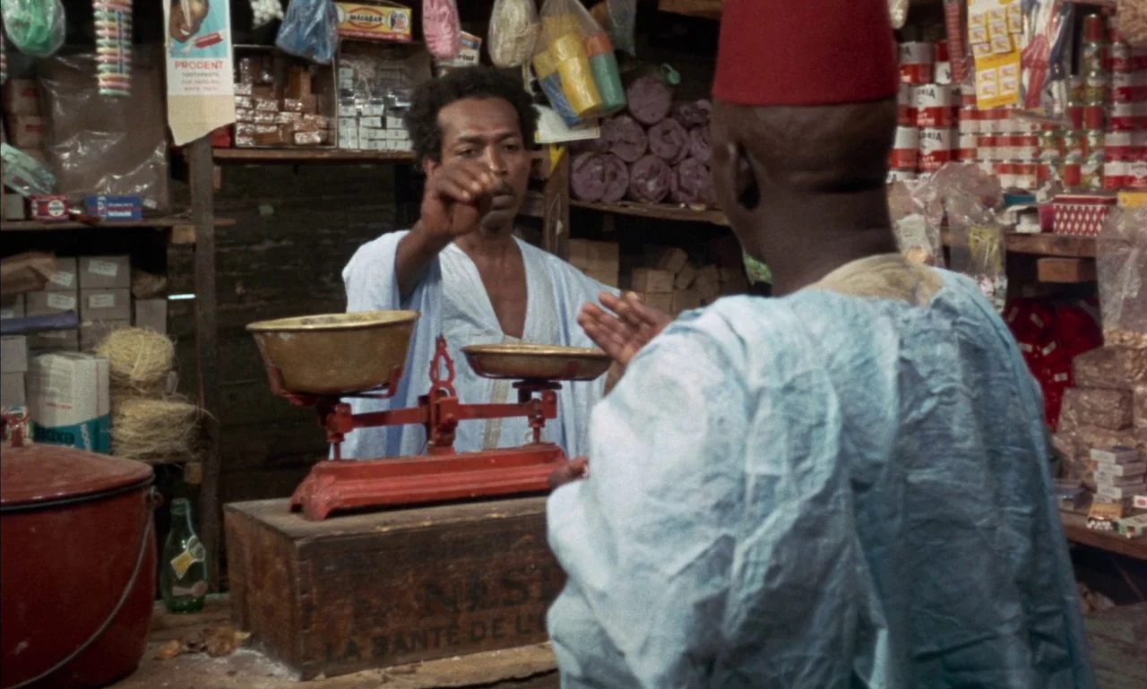 Mandabi (1968) movie still: medium — A cluttered shop interior features shelves stocked with various packaged goods and containers. A man interacts…