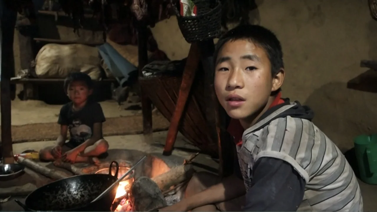 Hondar 2050 (2018) movie still: medium — A dark, earthen-floored room contains a central fire pit with various woven baskets overhead. Two boys sit…