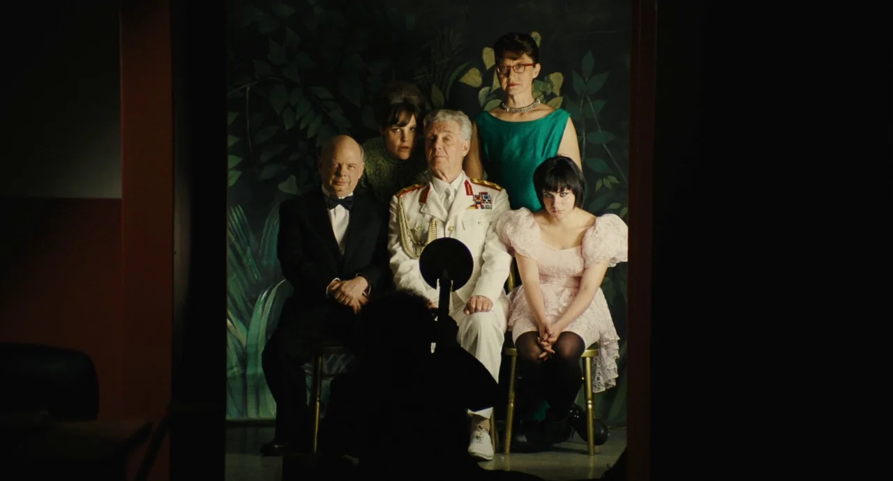 The Double (2013) movie still: medium — A formal family portrait is taken in a studio with a painted forest backdrop. Five people pose on chairs…