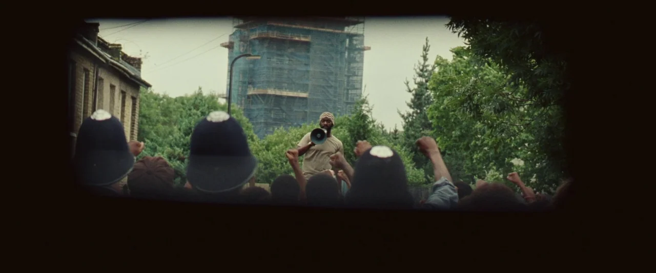 Mangrove (2020) movie still: wide — A group of protestors and uniformed police officers face a man with a megaphone. A large structure covered in…