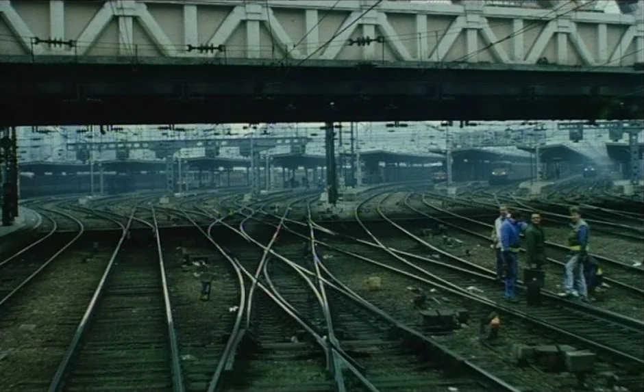 Simon, The Magician (1999) movie still: wide — A complex rail yard features multiple diverging metal tracks under a large concrete bridge. Four men…