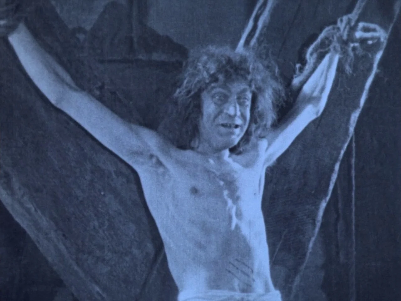 The Hunchback of Notre Dame (1923) movie still: medium — A man with wild hair is bound to a large wooden cross structure. He stands shirtless with…
