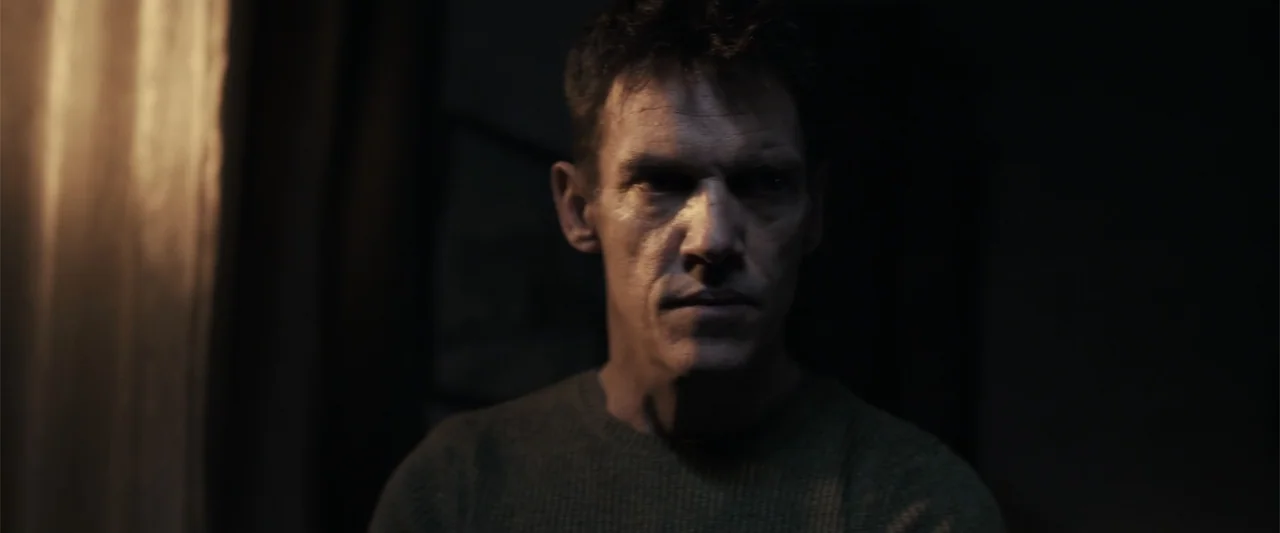 The Good Neighbor (2022) movie still: medium — A dimly lit interior space with a dark background. A middle-aged man in a sweater looks ahead while…