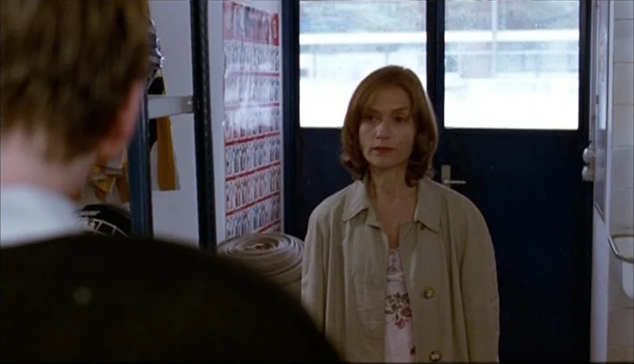 The Piano Teacher (2001) movie still: medium — A locker room with tiled walls and a large poster on the back wall. A woman in a trench coat stands…