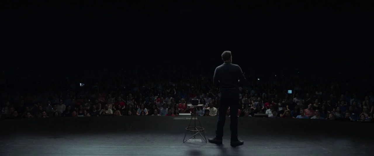 The Circle (2017) movie still: fullbody — A dark, expansive presentation hall is filled with a seated audience in shadow. A lone man stands on the…