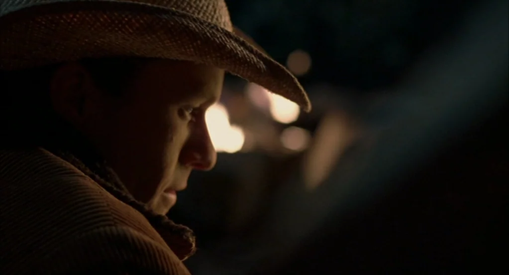Brokeback Mountain (2005) movie still: closeup — A man in a straw cowboy hat sits outdoors during the night. He looks toward the light of a fire…