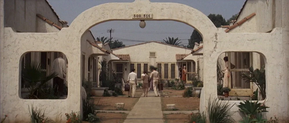The Fortune (1975) movie still: wide — A stucco-walled, U-shaped apartment complex with a central concrete path and a decorative archway. Three…