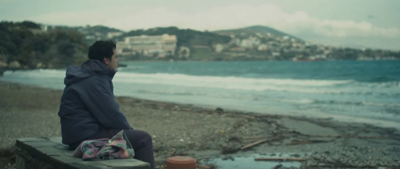 Alps (2011) movie still: medium — A pebble-strewn coastline extends toward a distant town at the base of low hills. A young man sits alone on a low…