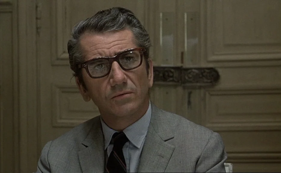 Murmur of the Heart (1971) movie still: closeup — A man in a suit sits before a closed wooden door with simple molding. He gazes off-screen while…