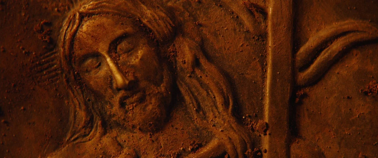 Silence (2016) movie still: closeup — A terracotta-toned relief sculpture depicts a stylized face in low-relief carving. Debris and dirt particles…