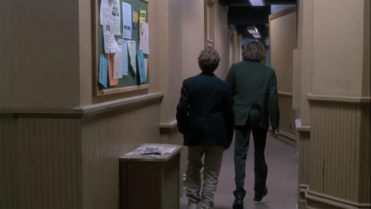 Boys (2021) movie still: fullbody — An institutional corridor features tan walls with beadboard paneling and a large bulletin board. Two teenagers…