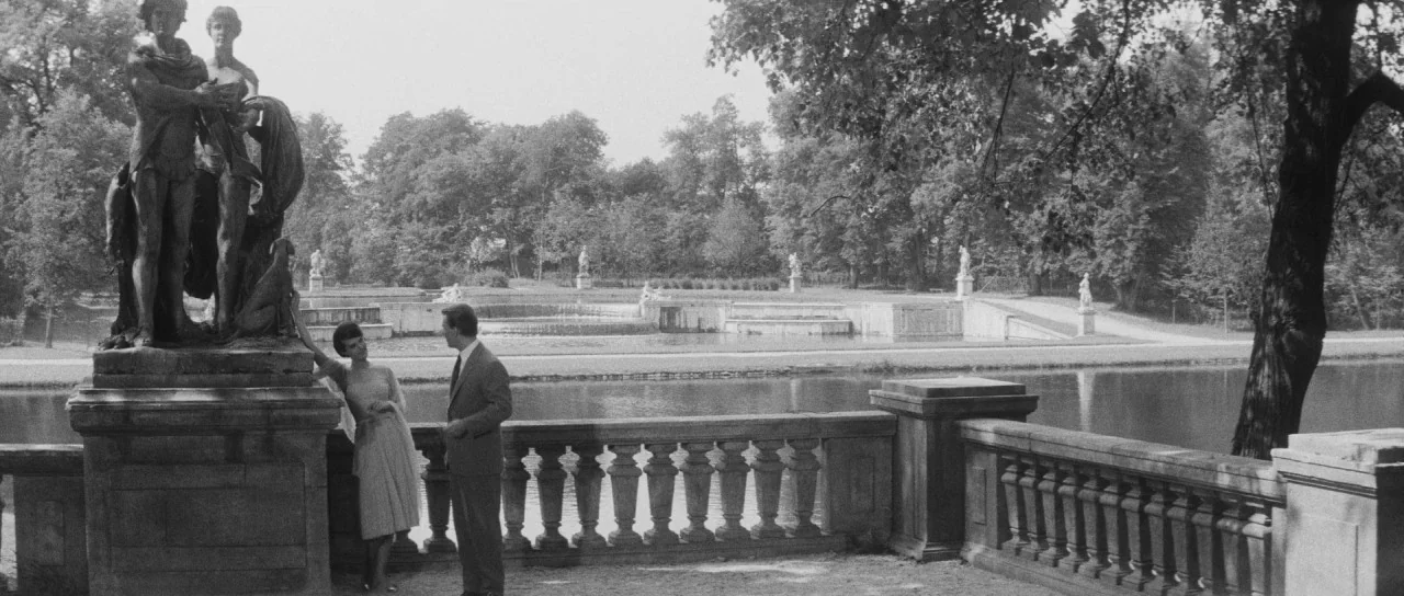 Last Year at Marienbad (1961) movie still: wide — A formal park setting features a stone balustrade and a large bronze sculptural group. A man and…