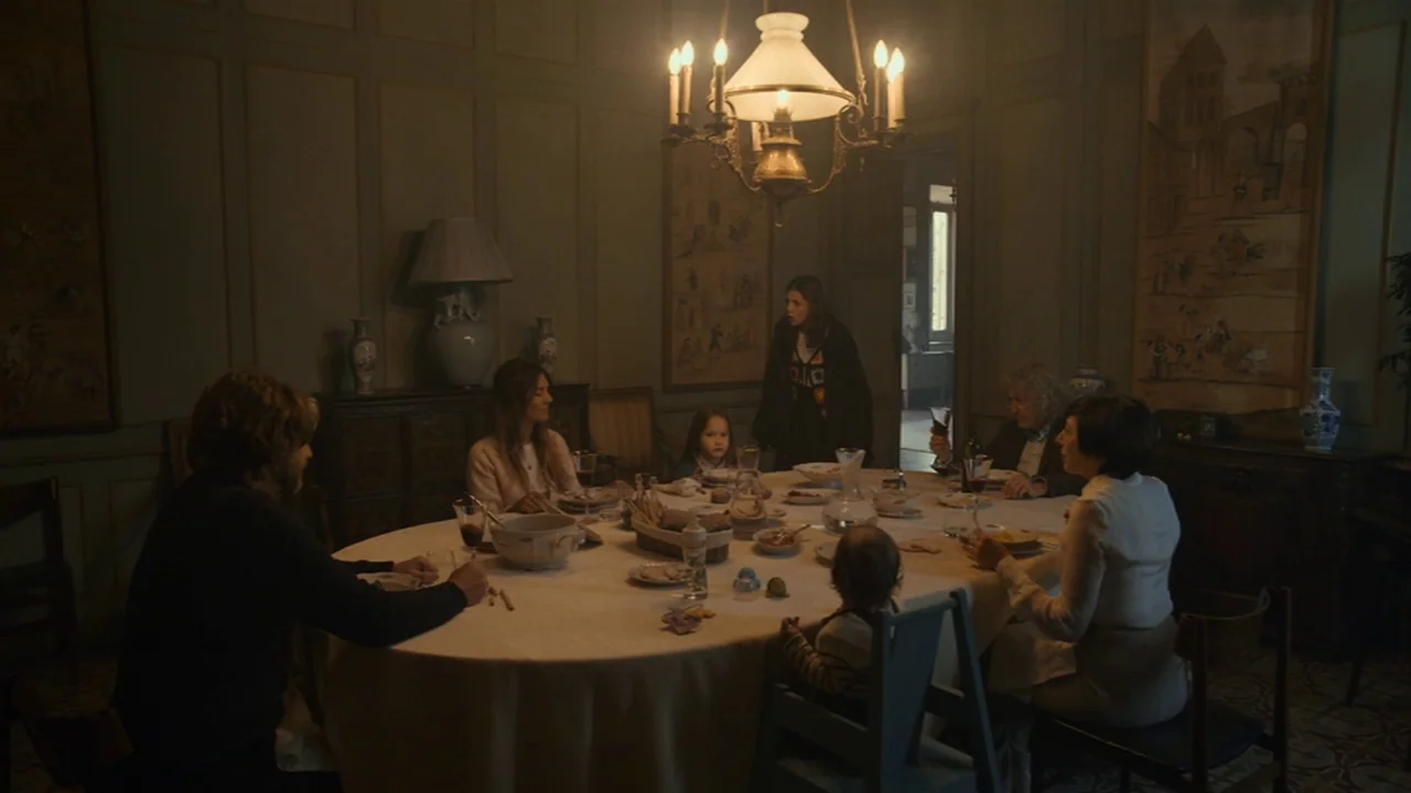 Amanda (2022) movie still: wide — A dimly lit room with paneled walls features a large circular table set for dining. A family is gathered around…