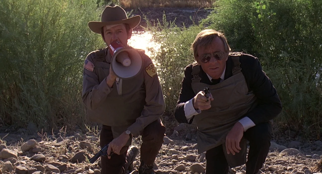 Charley Varrick (1973) movie still: medium — Two law enforcement officers in tactical gear kneel on a dry, rocky riverbank outdoors. One officer…