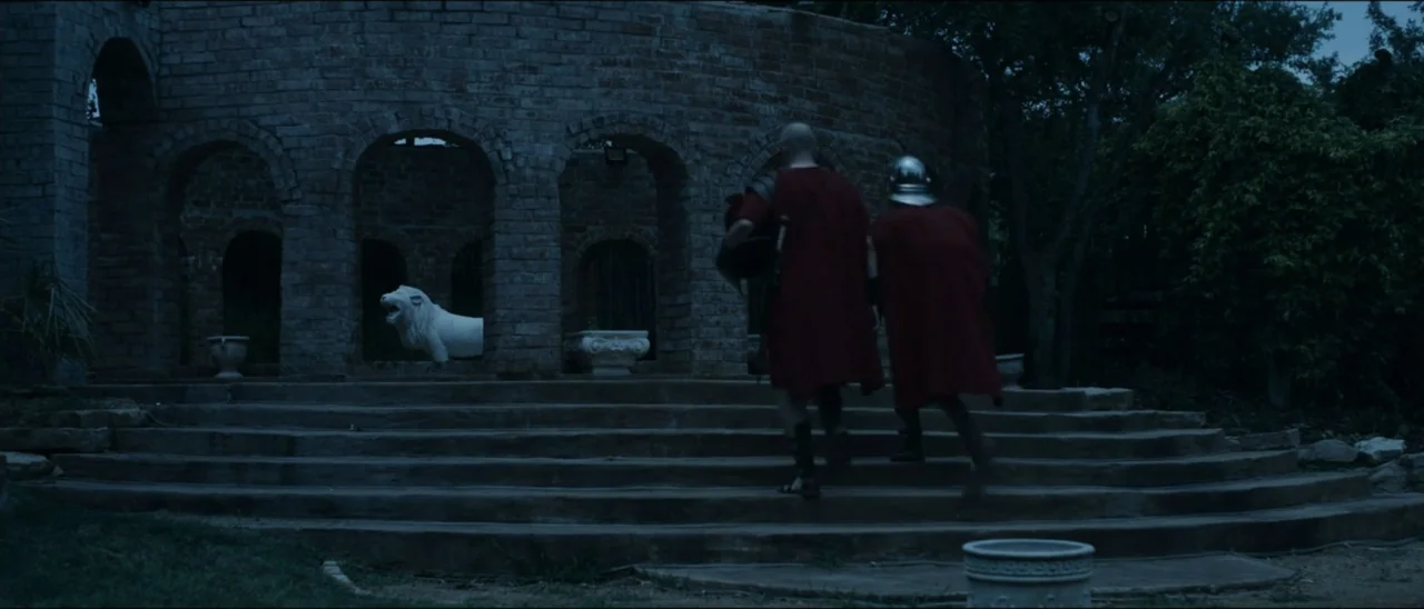 The Penitent Thief (2021) movie still: wide — A stone structure features tiered steps and arched bays containing white sculptures. Two men in…