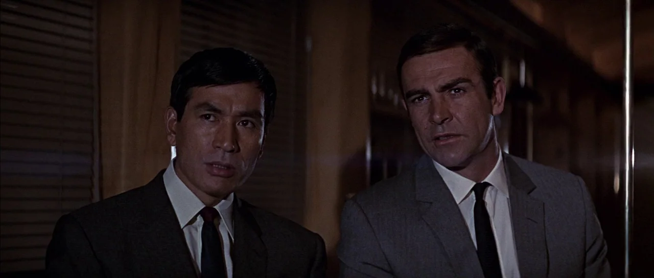 You Only Live Twice (1967) movie still: medium — A dimly lit indoor space features vertical window blinds covered by heavy curtains behind subjects.…