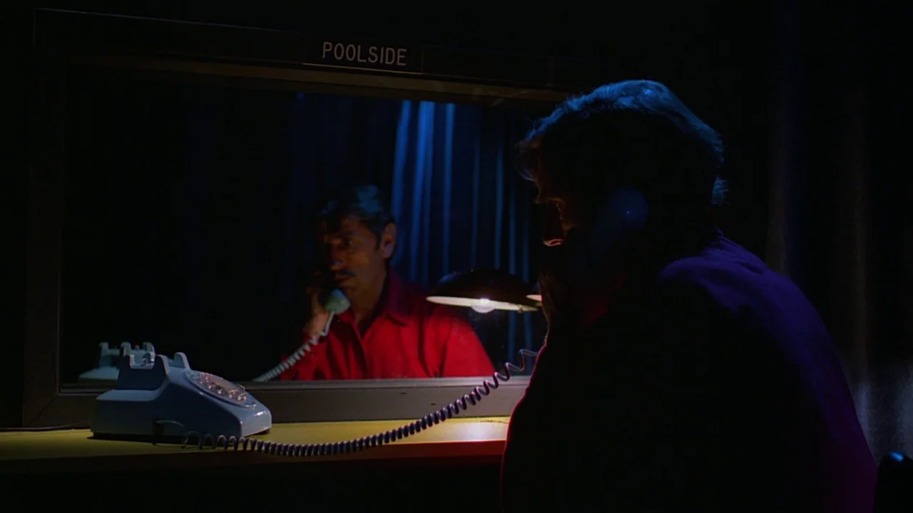 Paris, Texas (1984) movie still: medium — A dark, cramped space features a reflective glass panel labeled Poolside above a desk. A man with a…