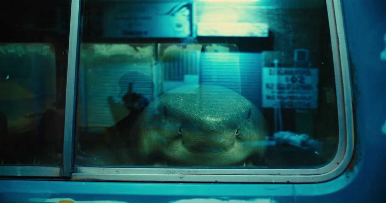 The Suicide Squad (2021) movie still: closeup — A shark sits inside a dark water tank viewed through a rectangular glass window. The creature faces…