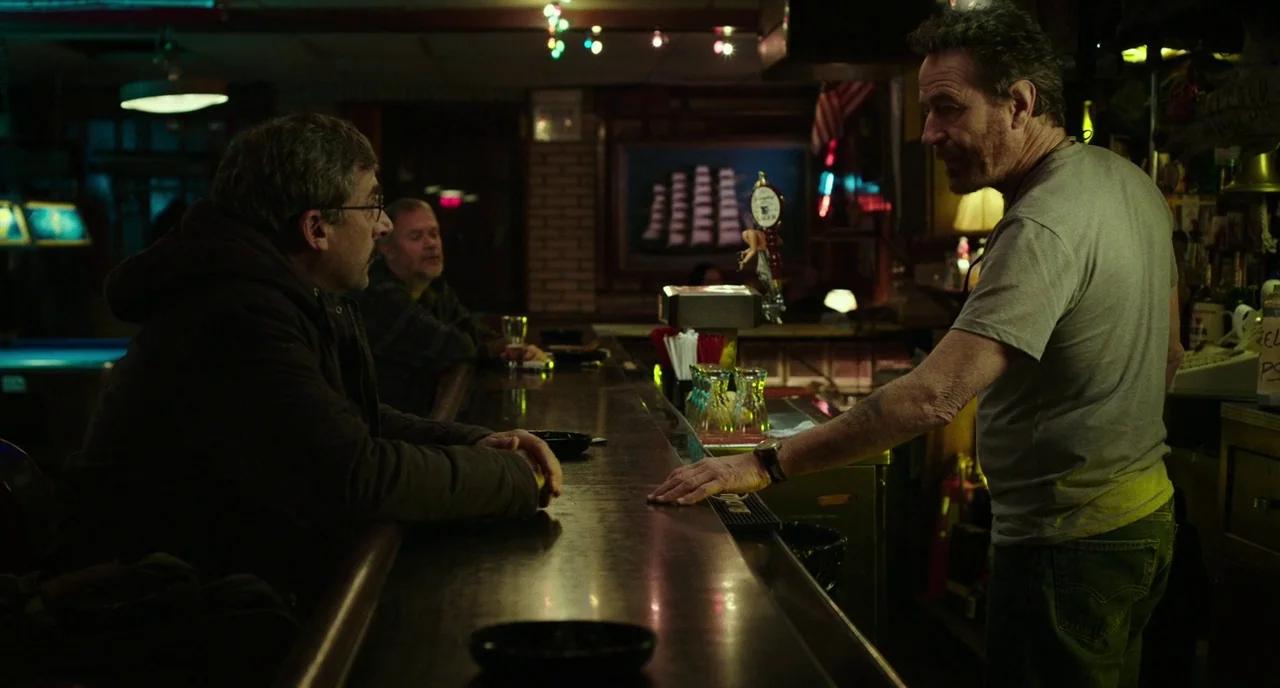 Last Flag Flying (2017) movie still: medium — A dimly lit local tavern featuring a long wooden counter and cluttered shelves. A man in a grey…