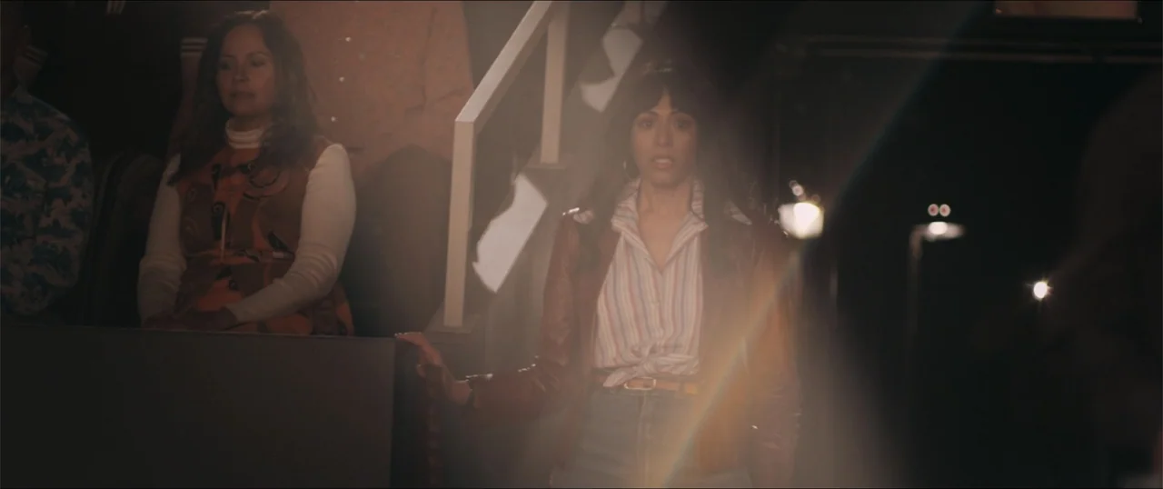Woman of the Hour (2024) movie still: medium — An interior space features tiered seating and a dark partition in the foreground. A young woman with…