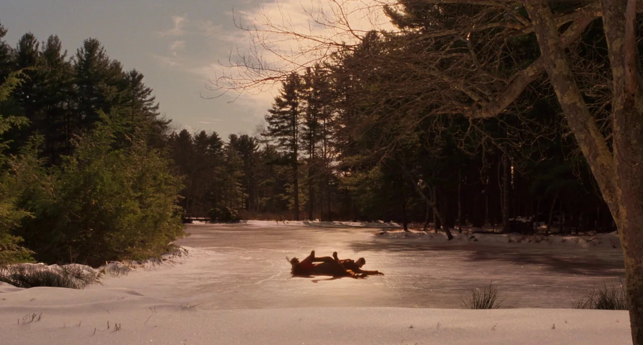 Little Women (2019) movie still: wide — A frozen pond is bordered by a dense forest under a winter sky. Two figures lie together on the ice in the…