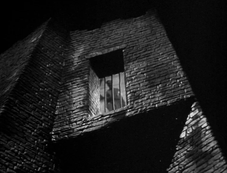 Frankenstein movie still: establishing — A stone tower built from dark bricks rises into the dark night sky. A singular window with vertical iron…
