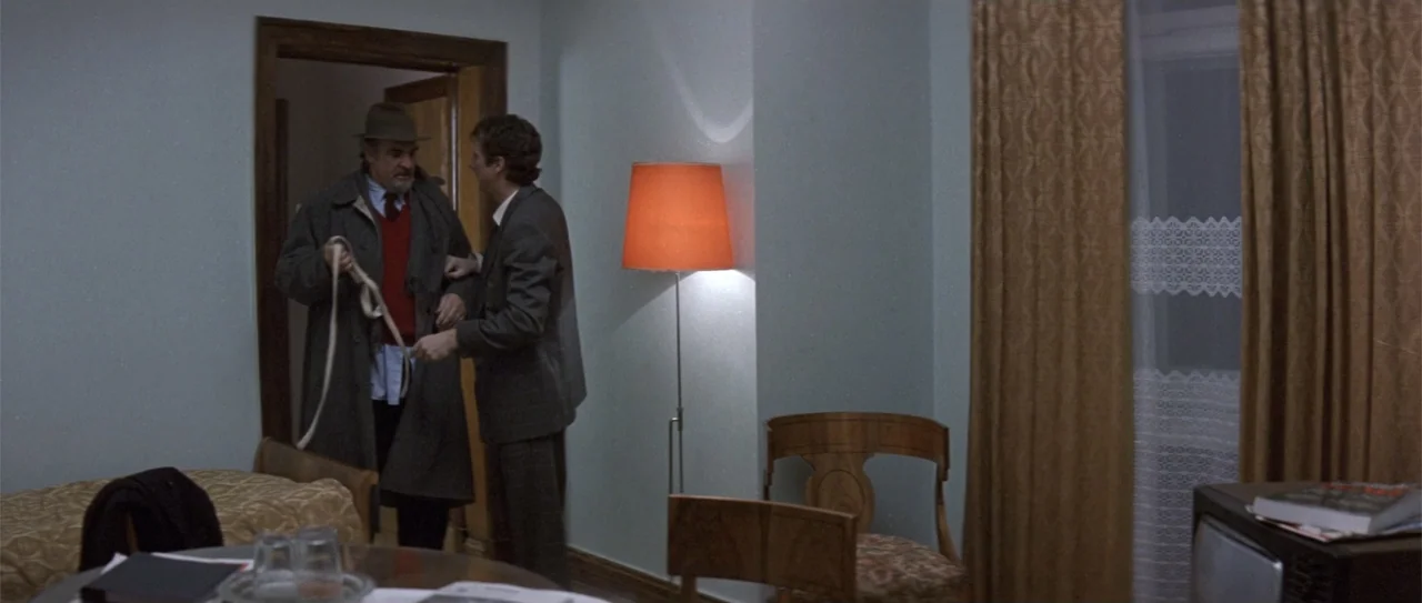 The Russia House (1990) movie still: medium — An interior hotel room features pale walls, a standing orange lamp, and wooden furniture. Two men…