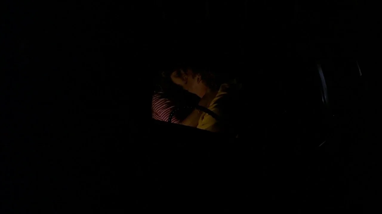The Slumber Party Massacre (1982) movie still: medium — A dark vehicle interior with very limited visibility beyond the two people sitting inside.…