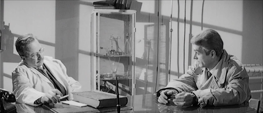 I Vampiri (1957) movie still: medium — An interior office with a glass cabinet and a desk in the center. Two men sit opposite one another at the…