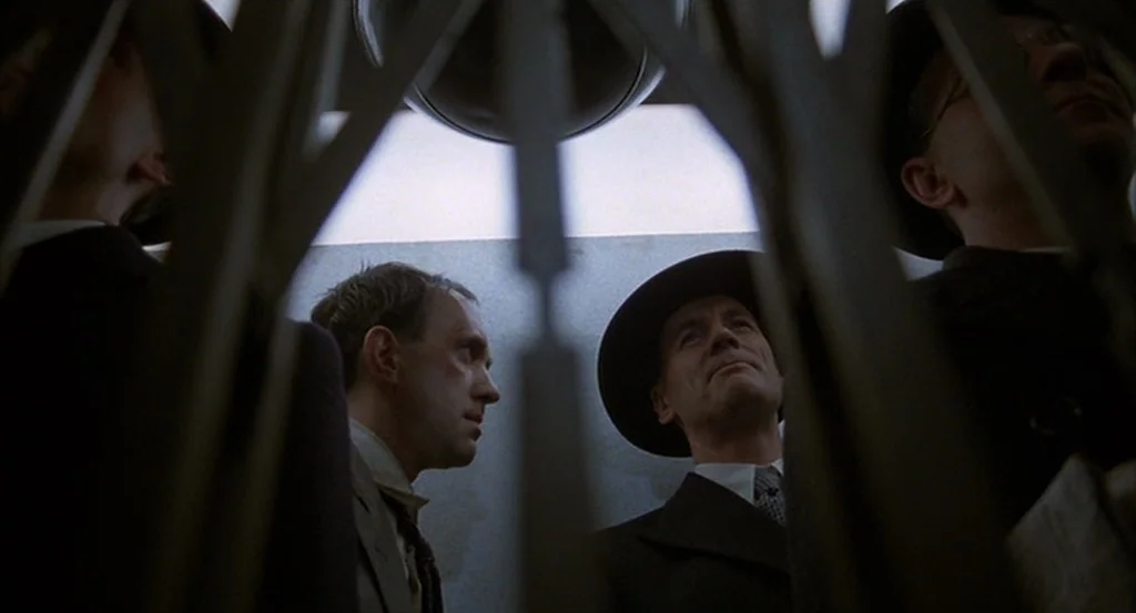 Brazil (1985) movie still: medium — An interior shot looking up through an open metal concertina gate of an elevator. Several men in dark suits and…