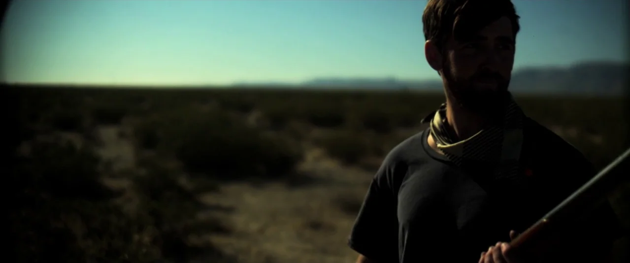 Bellflower (2011) movie still: medium — A man stands in a vast, arid desert under a bright blue sky. The man holds an unidentified object while…