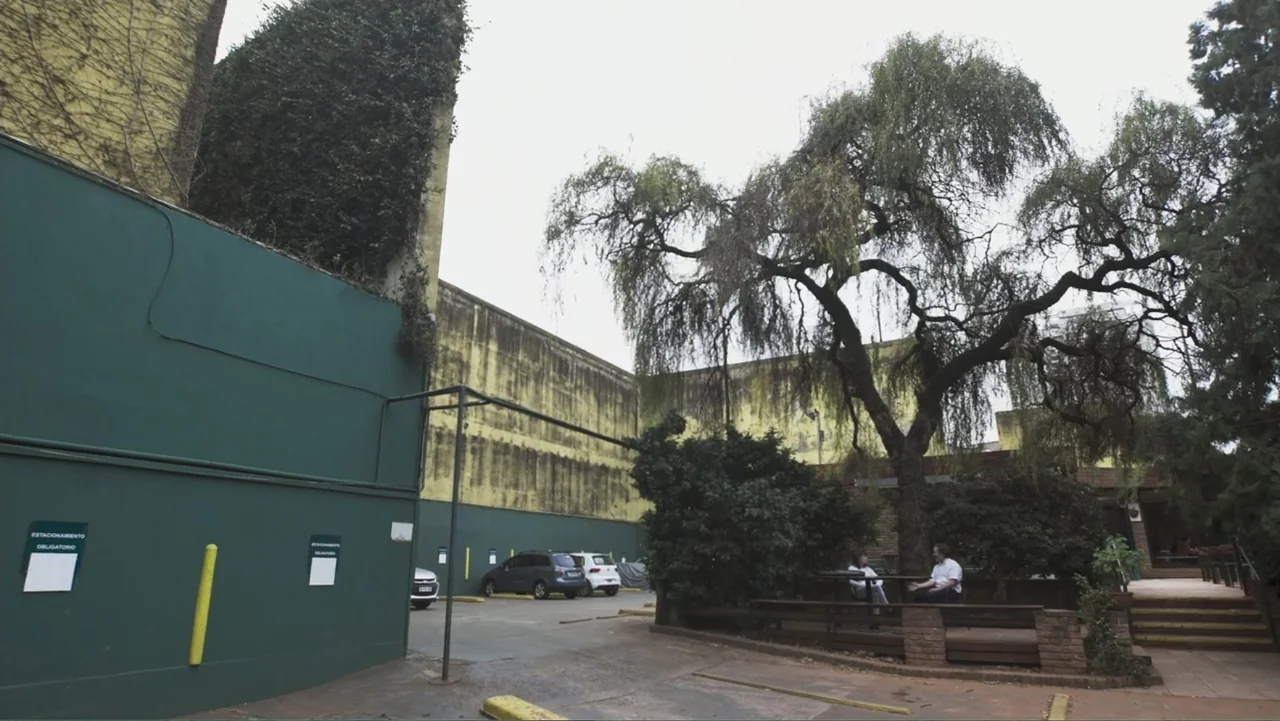 Smog in Your Heart (2022) movie still: wide — A paved parking lot features a high green wall with an overgrown vine covering. Two individuals sit at…