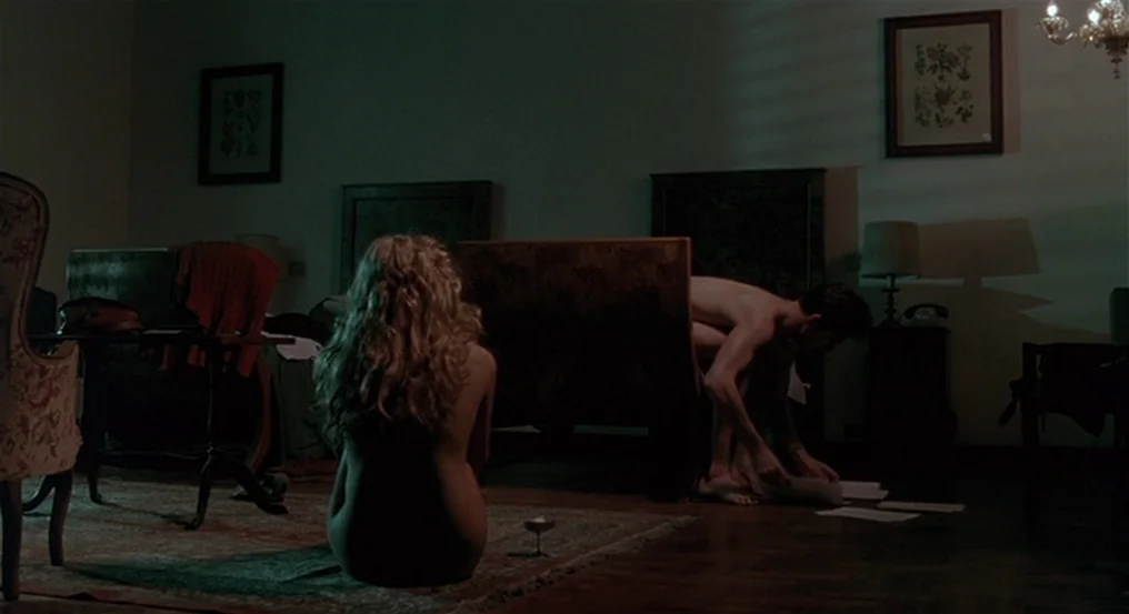 The Comfort of Strangers (1990) movie still: wide — A dimly lit room contains patterned armchairs and a small rug on a dark floor. A blonde woman…