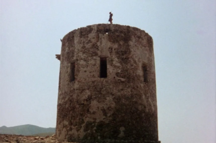 Sebastiane (1976) movie still: wide — A weathered, circular stone tower stands against an overcast, light grey sky. A small figure stands atop the…
