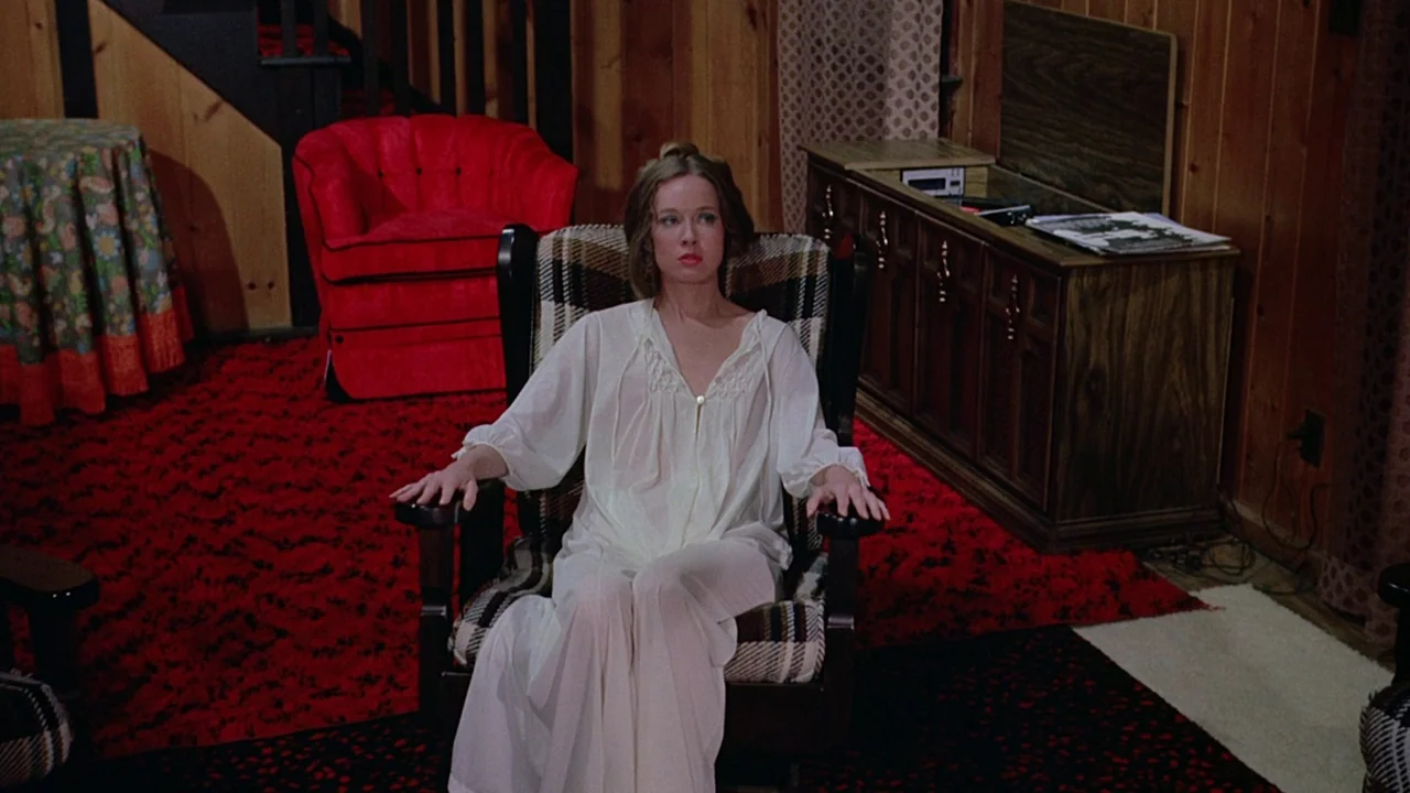 I Spit On Your Grave (1978) movie still: medium — A 1970s living room features wood-paneled walls and a bold red patterned carpet. A woman in a…