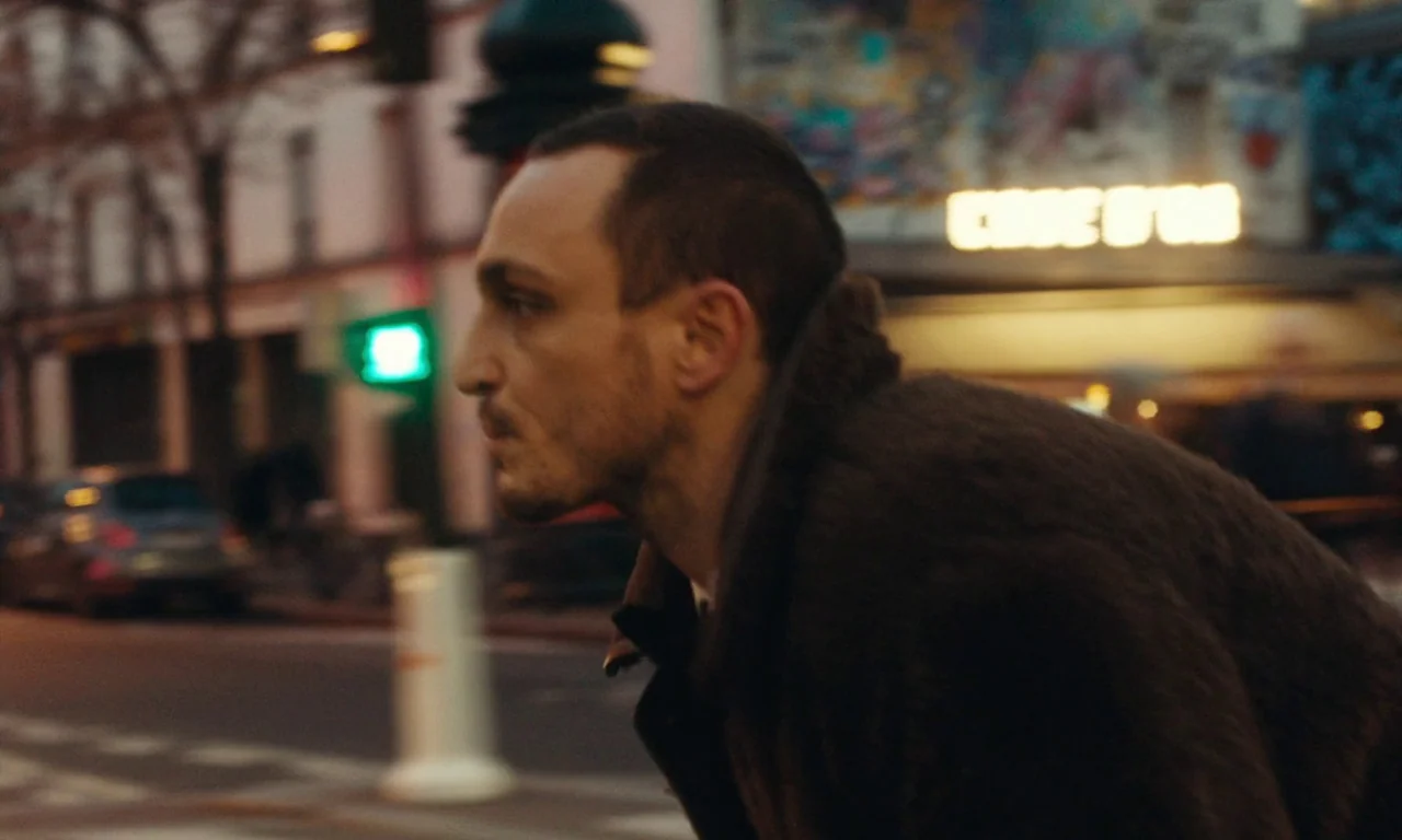 Passages (2023) movie still: medium — A bustling city street at night features blurred lights and evening traffic. A young man with a short haircut…