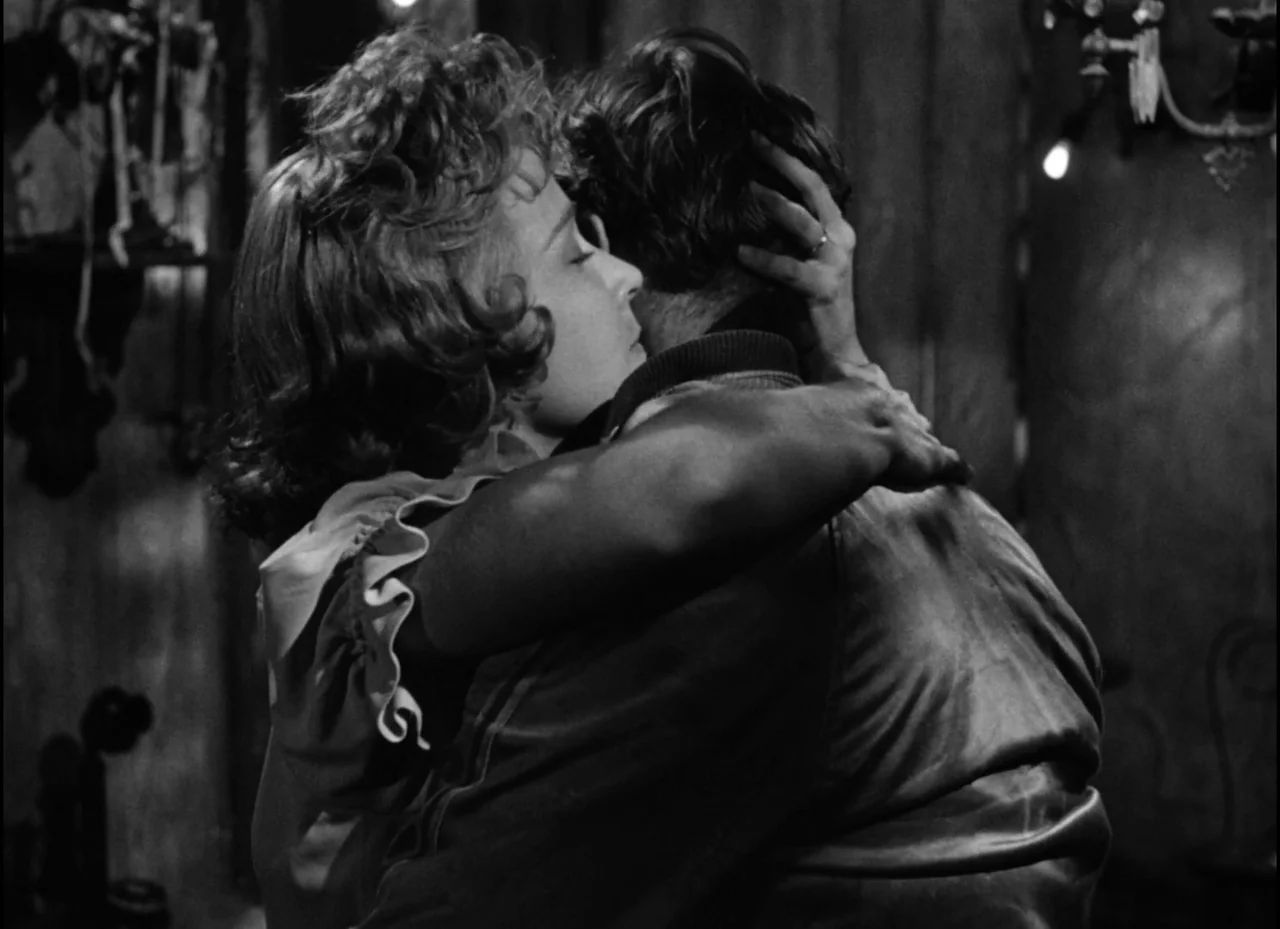 A Streetcar Named Desire (1951) movie still: medium — A dimly lit indoor space with textured walls and an ornate wall light fixture. A woman with…