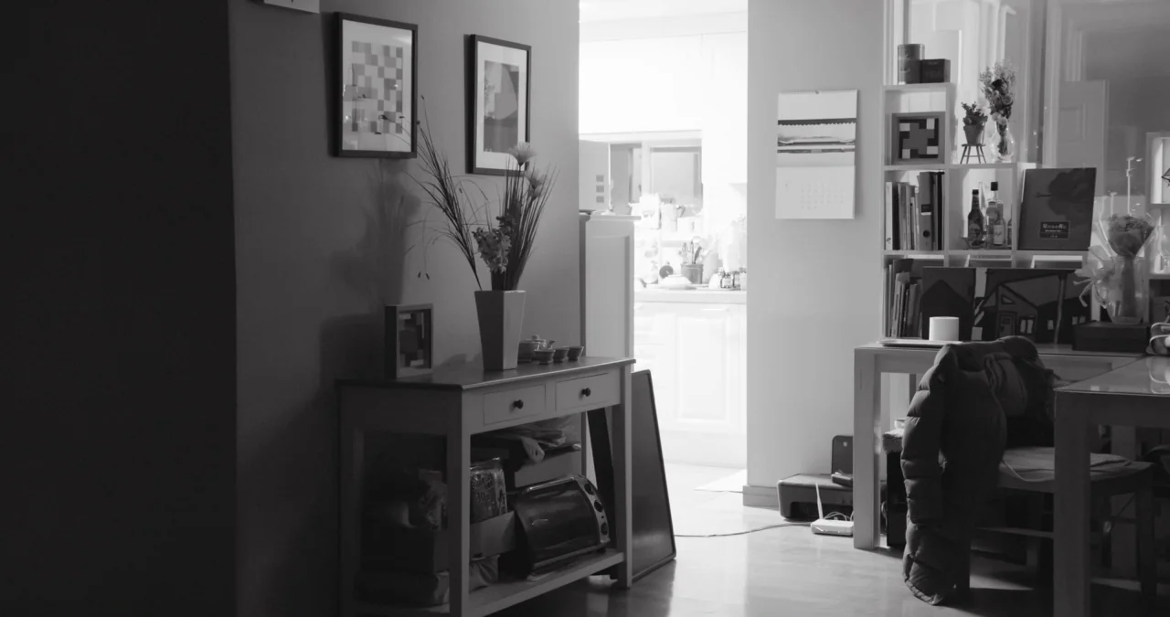 The Day After movie still: wide — A living area features a wooden console table topped with a vase and framed pictures. A bookshelf stands beside a…