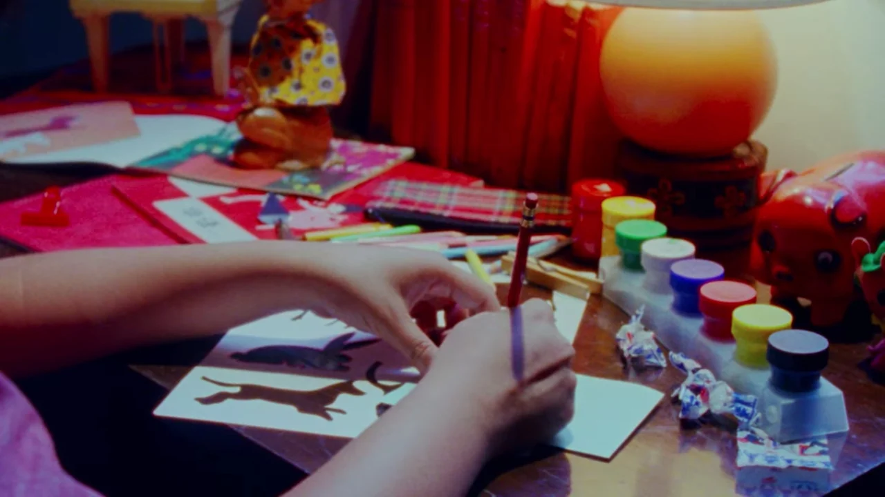 Shirkers (2018) movie still: closeup — A cluttered desk features a variety of art supplies and small plastic figurines. Hands are seen in the…