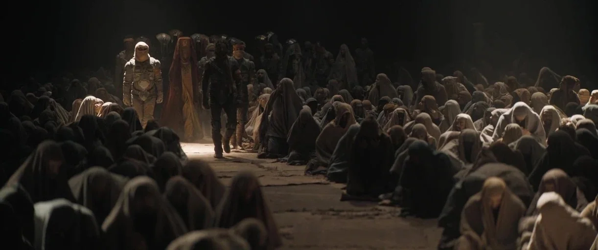 Dune: Part 2 (2024) movie still: wide — A vast underground cavern houses a dense crowd of figures wearing tattered hooded cloaks. Two central…