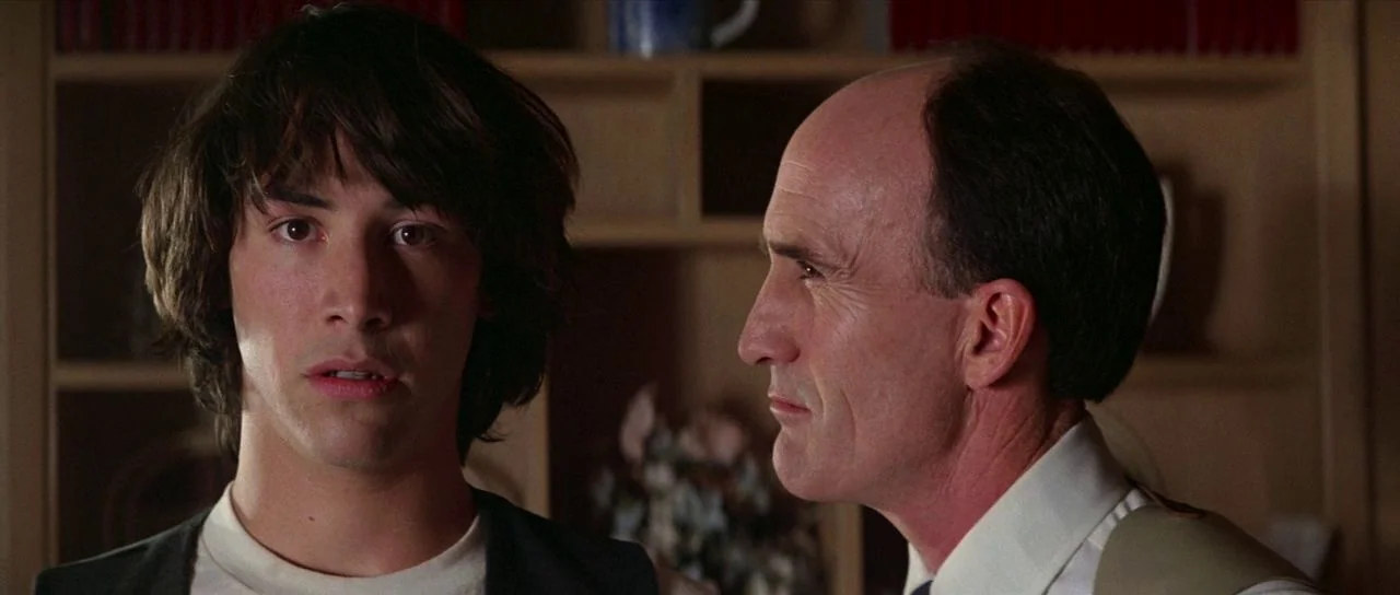 Bill and Ted’s Excellent Adventure (1989) movie still: medium — A domestic space features wooden shelving units filled with books in the background.…
